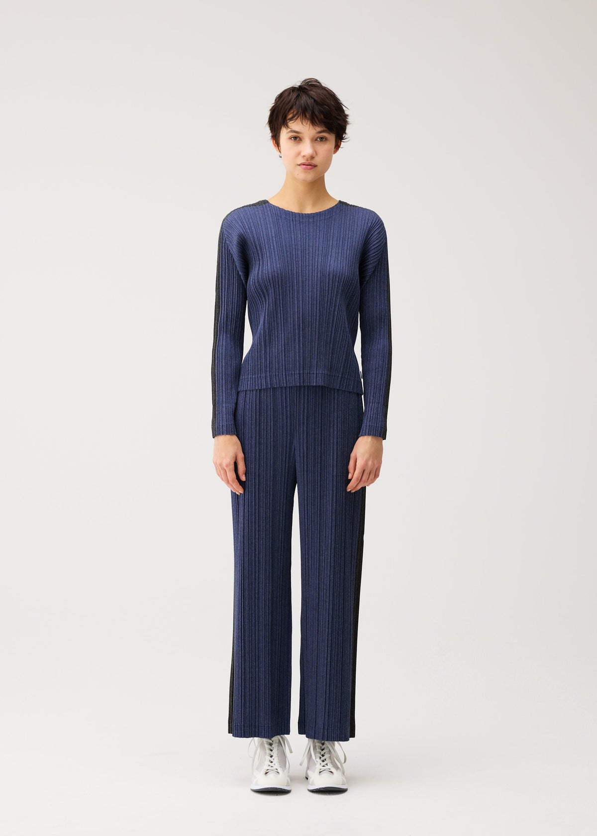 A person stands facing forward in the PLEATS PLEASE ISSEY MIYAKE COTTON DENIM set—matching blue long-sleeved top and wide-leg pants—paired with white sneakers, against a plain light background. view 5 of 8