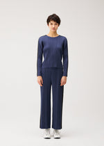 A person stands facing forward in the PLEATS PLEASE ISSEY MIYAKE COTTON DENIM set—matching blue long-sleeved top and wide-leg pants—paired with white sneakers, against a plain light background.