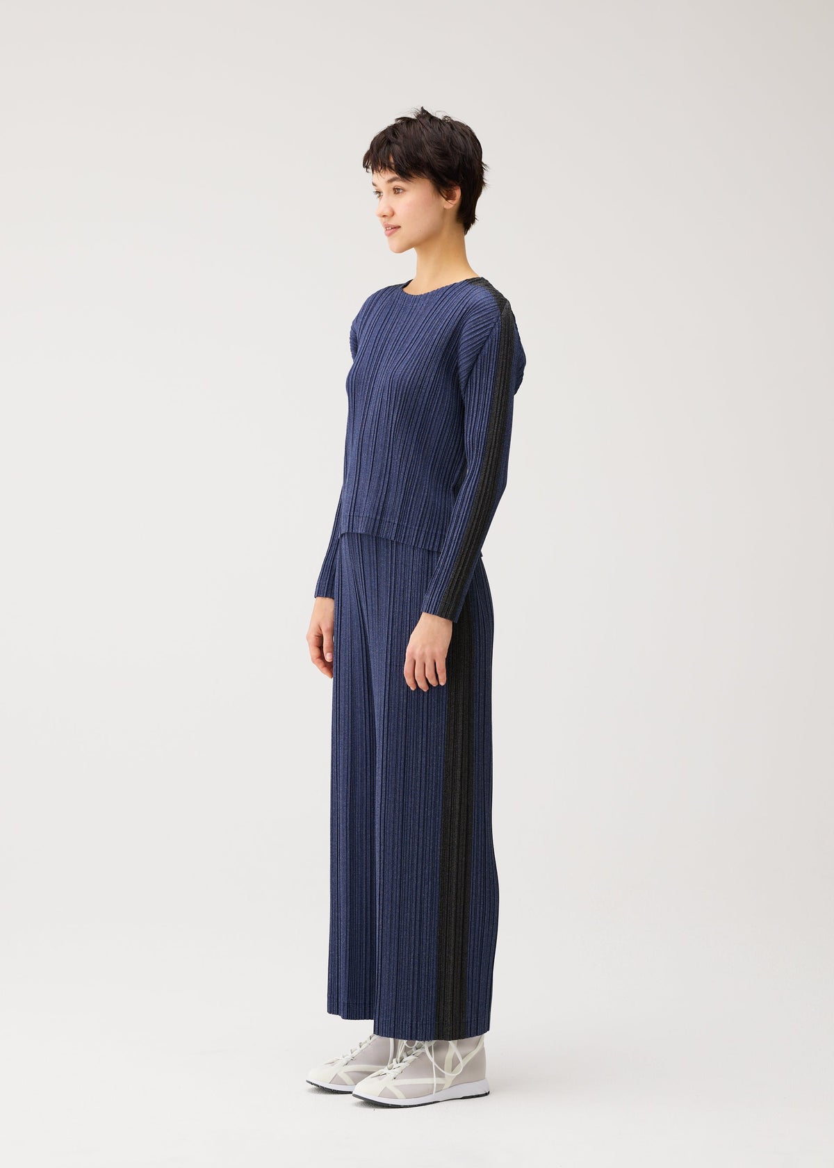 Zoomed view A woman in profile models the COTTON DENIM round-neck top and wide-leg pants by PLEATS PLEASE ISSEY MIYAKE with white lace-up shoes, set against a plain white background.
