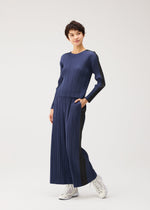 Model wears the COTTON DENIM set by PLEATS PLEASE ISSEY MIYAKE—ribbed navy long-sleeve top and wide-leg pants with hands in pockets, styled with white high-top sneakers against a plain white background.