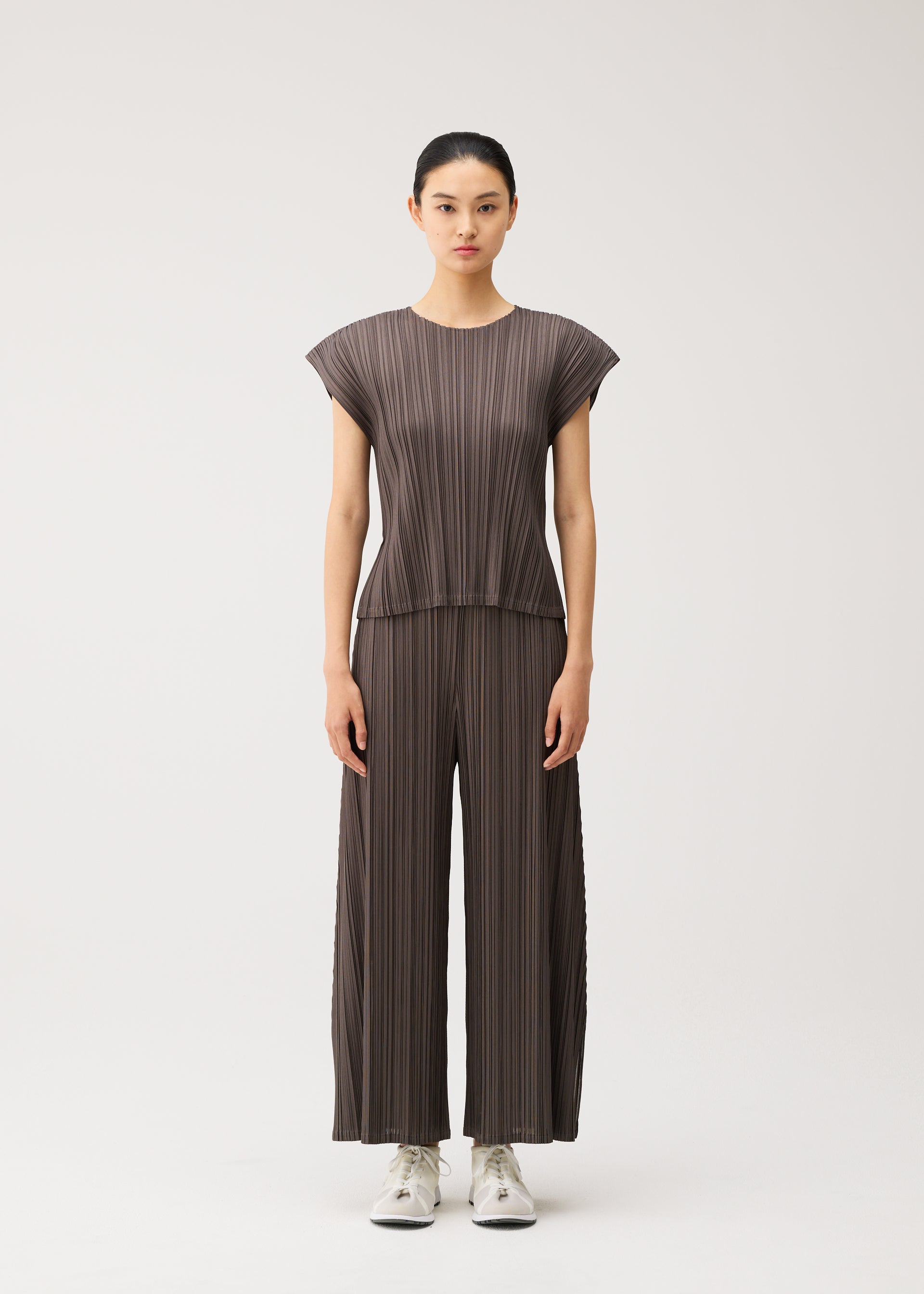 Zoomed view A woman stands facing forward against a plain light background, dressed in the MELLOW PLEATS set by PLEATS PLEASE ISSEY MIYAKE—a loose-fitting, pleated brown round-neck top with French sleeves and matching wide-leg pants made from soft-textured fabric—paired with white sneakers.