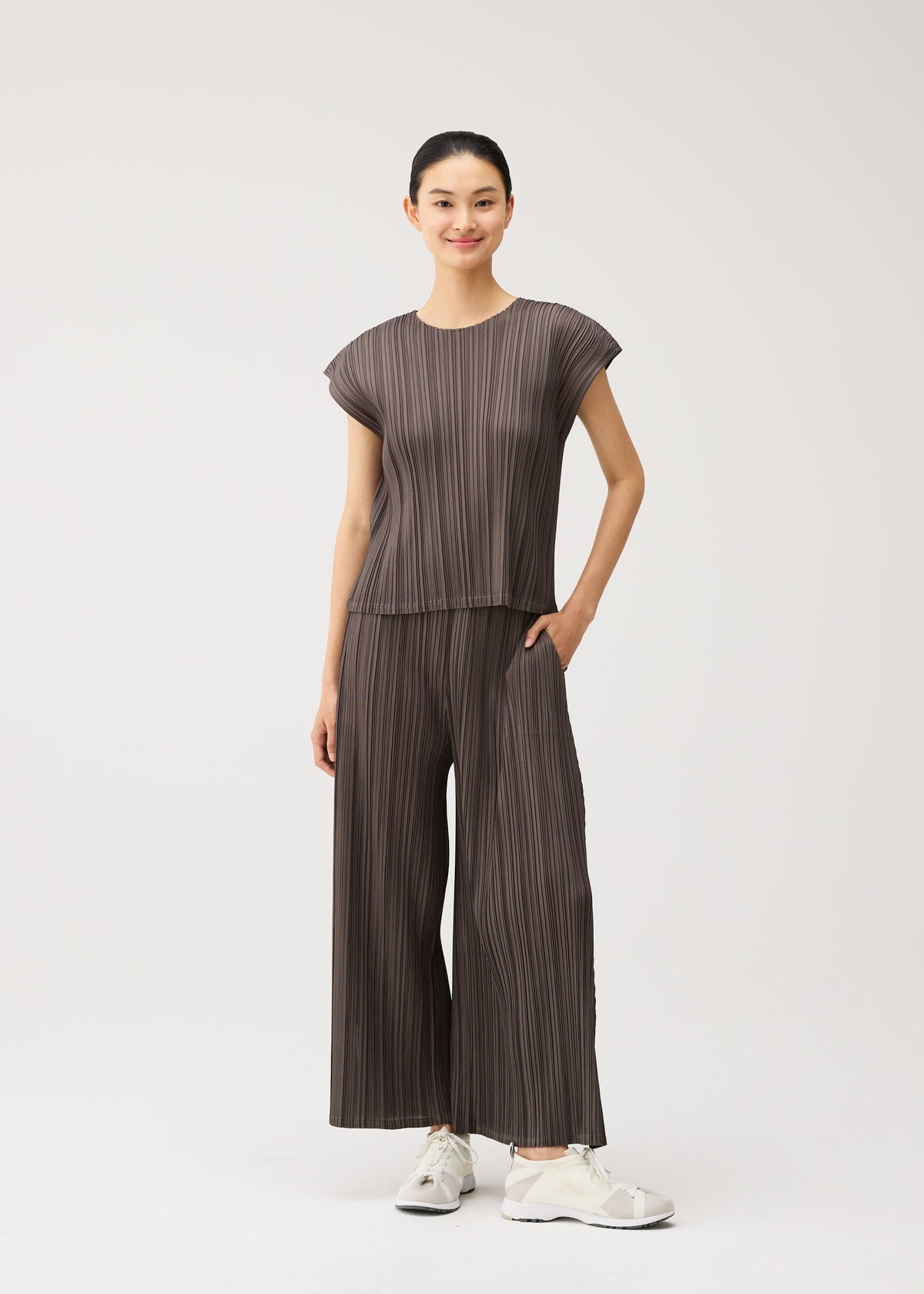 A woman stands smiling against a plain white background, wearing the MELLOW PLEATS set from PLEATS PLEASE ISSEY MIYAKE—a loose, pleated brown round-neck top with French sleeves and matching wide-legged pants crafted from soft-textured fabric. She pairs the outfit with white sneakers and has one hand in her pocket. view 8 of 8