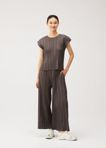 A woman stands smiling against a plain white background, wearing the MELLOW PLEATS set from PLEATS PLEASE ISSEY MIYAKE—a loose, pleated brown round-neck top with French sleeves and matching wide-legged pants crafted from soft-textured fabric. She pairs the outfit with white sneakers and has one hand in her pocket.