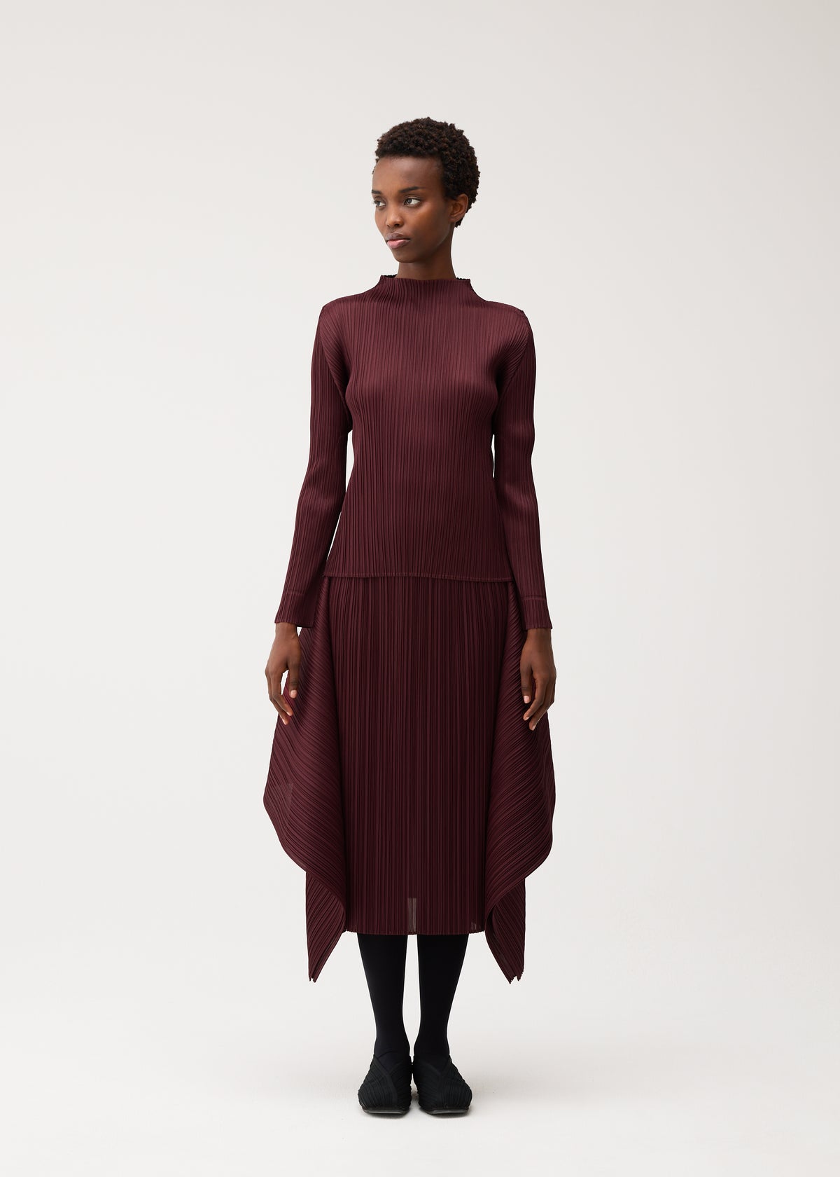 A woman stands against a light background, modeling the FLUTTER dress by PLEATS PLEASE ISSEY MIYAKE—a long-sleeved, burgundy pleated piece featuring an asymmetrical hem and high-neck detail. She finishes the ensemble with black tights and black shoes. view 4 of 7
