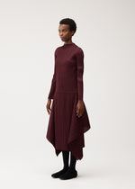 A person stands against a plain background, showcasing women’s fashion in the FLUTTER long-sleeved, burgundy pleated dress with an asymmetrical hem from PLEATS PLEASE ISSEY MIYAKE, paired with black tights and black shoes.