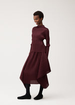 A person smiles while wearing the FLUTTER set by PLEATS PLEASE ISSEY MIYAKE—a burgundy, long-sleeved pleated top and matching asymmetrical skirt—paired with black tights and shoes, standing against a plain light background for a chic look that highlights timeless women’s fashion.