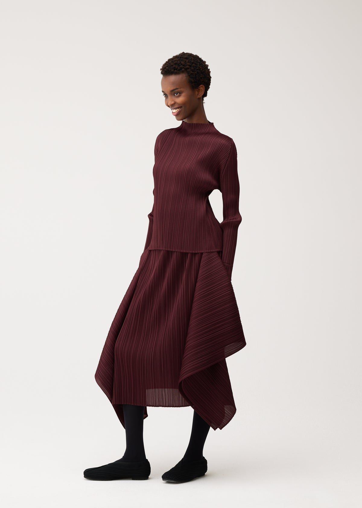 Zoomed view A person smiles while wearing the FLUTTER set by PLEATS PLEASE ISSEY MIYAKE—a burgundy, long-sleeved pleated top and matching asymmetrical skirt—paired with black tights and shoes, standing against a plain light background for a chic look that highlights timeless women’s fashion.