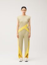 A woman stands against a plain background wearing the TWIRL sleeveless, round-neck pleated jumpsuit in a beige and yellow geometric pattern from PLEATS PLEASE ISSEY MIYAKE, paired with white sneakers. She looks directly at the camera with a neutral expression.