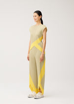 A woman stands sideways, smiling, wearing the TWIRL beige pleated dress with yellow geometric patterns and a round-neck sleeveless top by PLEATS PLEASE ISSEY MIYAKE, paired with white sneakers, against a plain light background.