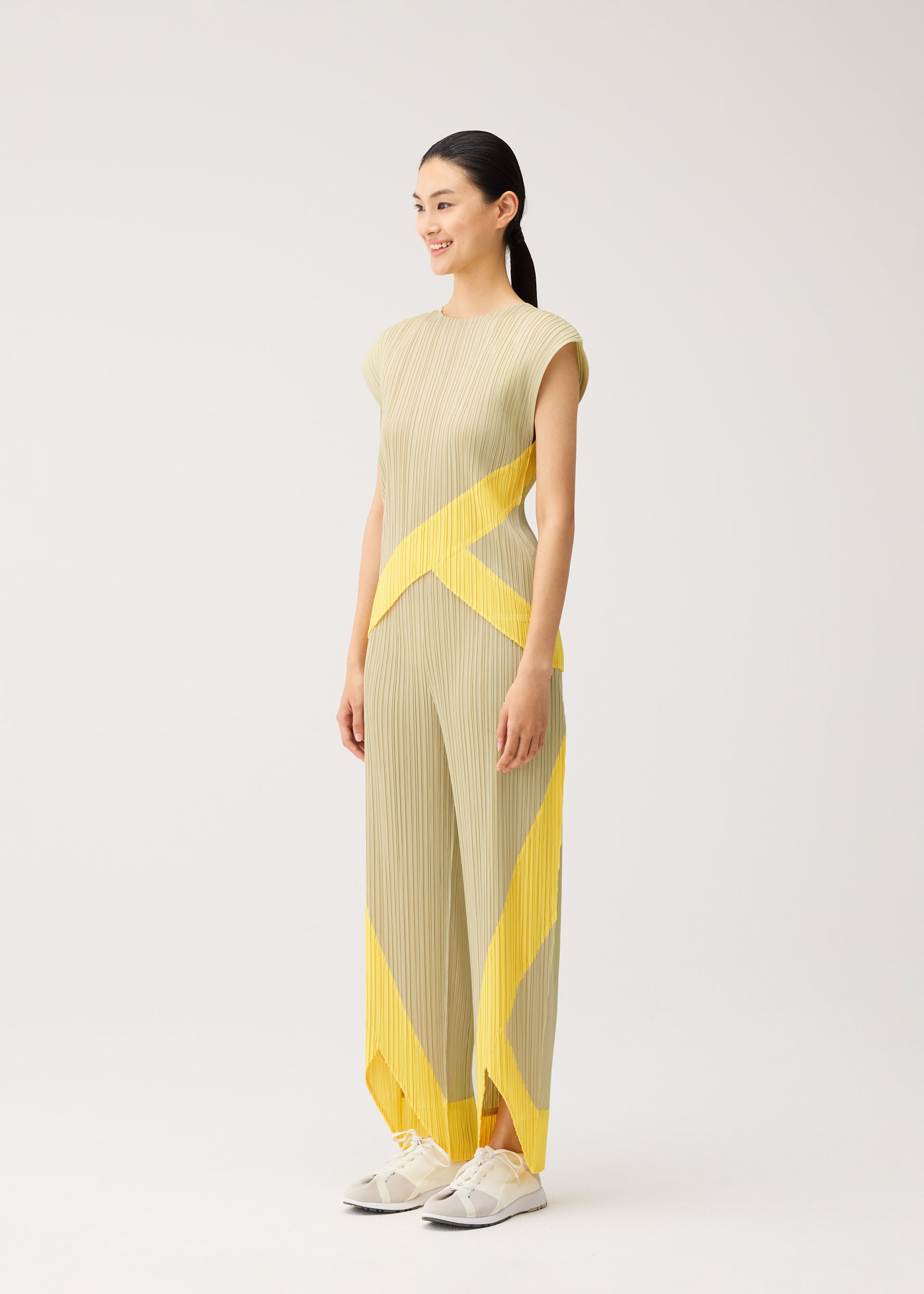 Zoomed view A woman stands sideways, smiling, wearing the TWIRL beige pleated dress with yellow geometric patterns and a round-neck sleeveless top by PLEATS PLEASE ISSEY MIYAKE, paired with white sneakers, against a plain light background.