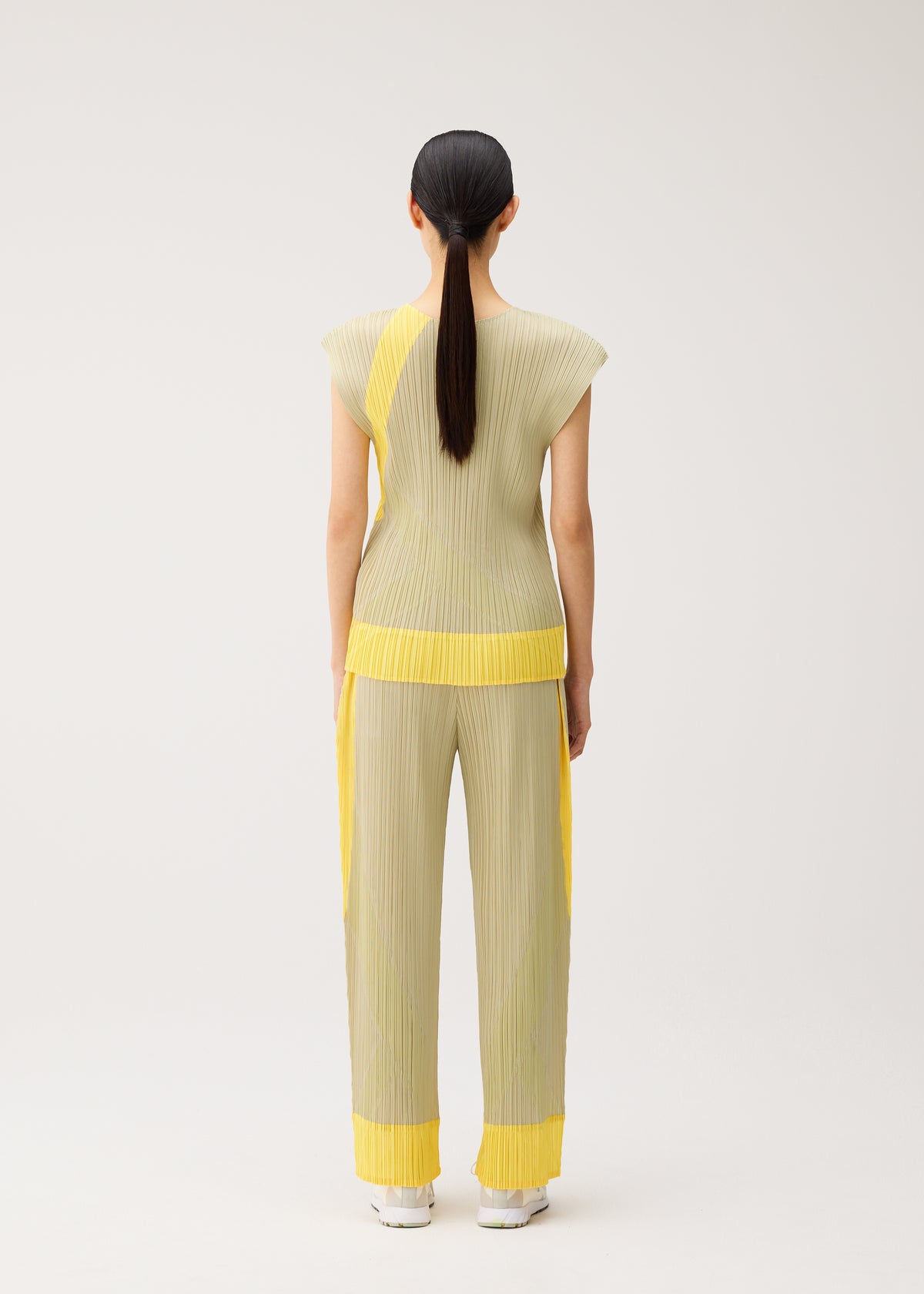 Model with long, dark hair in a ponytail faces away from the camera, wearing the TWIRL set by PLEATS PLEASE ISSEY MIYAKE—a light yellow, round-neck sleeveless top and pants featuring bright yellow accents—posed in a minimal studio setting. view 7 of 9