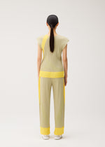Model with long, dark hair in a ponytail faces away from the camera, wearing the TWIRL set by PLEATS PLEASE ISSEY MIYAKE—a light yellow, round-neck sleeveless top and pants featuring bright yellow accents—posed in a minimal studio setting.