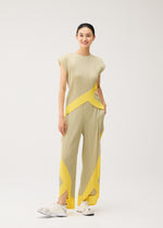 A woman stands smiling in PLEATS PLEASE ISSEY MIYAKE's TWIRL ensemble—a pleated beige outfit with yellow geometric accents, featuring a sleeveless round-neck top. Her hair is tied back and she pairs the look with white sneakers, set against a plain light background.