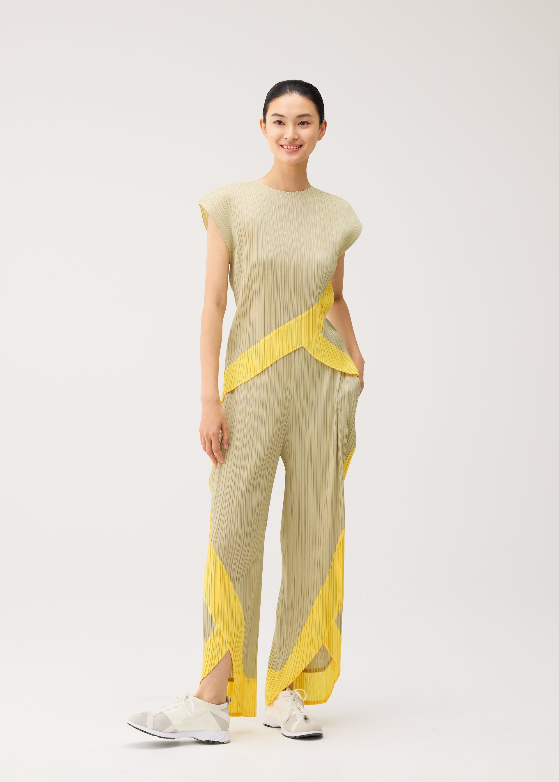 Zoomed view A woman stands smiling in PLEATS PLEASE ISSEY MIYAKE's TWIRL ensemble—a pleated beige outfit with yellow geometric accents, featuring a sleeveless round-neck top. Her hair is tied back and she pairs the look with white sneakers, set against a plain light background.
