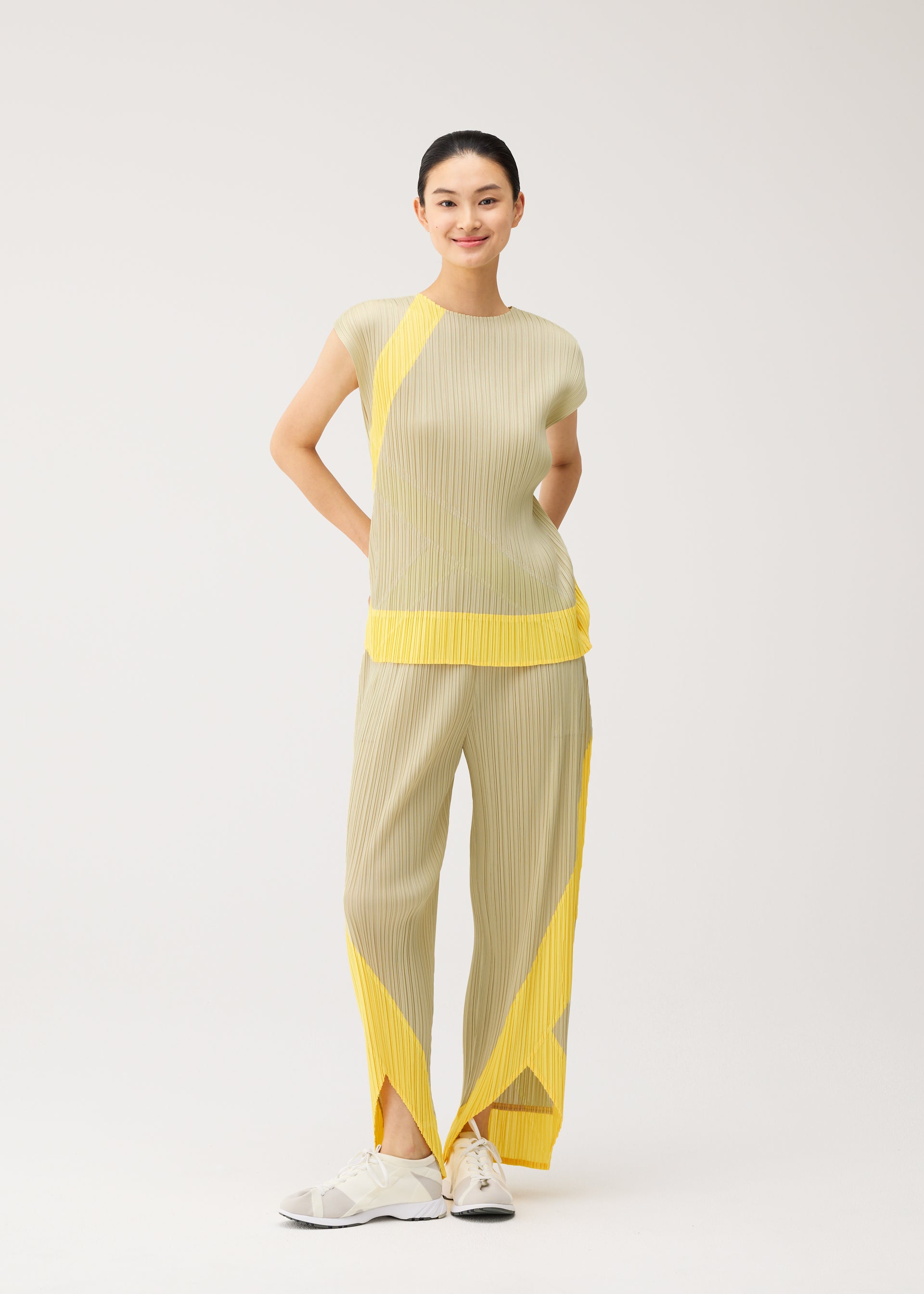Zoomed view A woman stands smiling against a plain light background, wearing the TWIRL set from PLEATS PLEASE ISSEY MIYAKE—a beige pleated sleeveless top and pants with yellow geometric accents—paired with white sneakers.