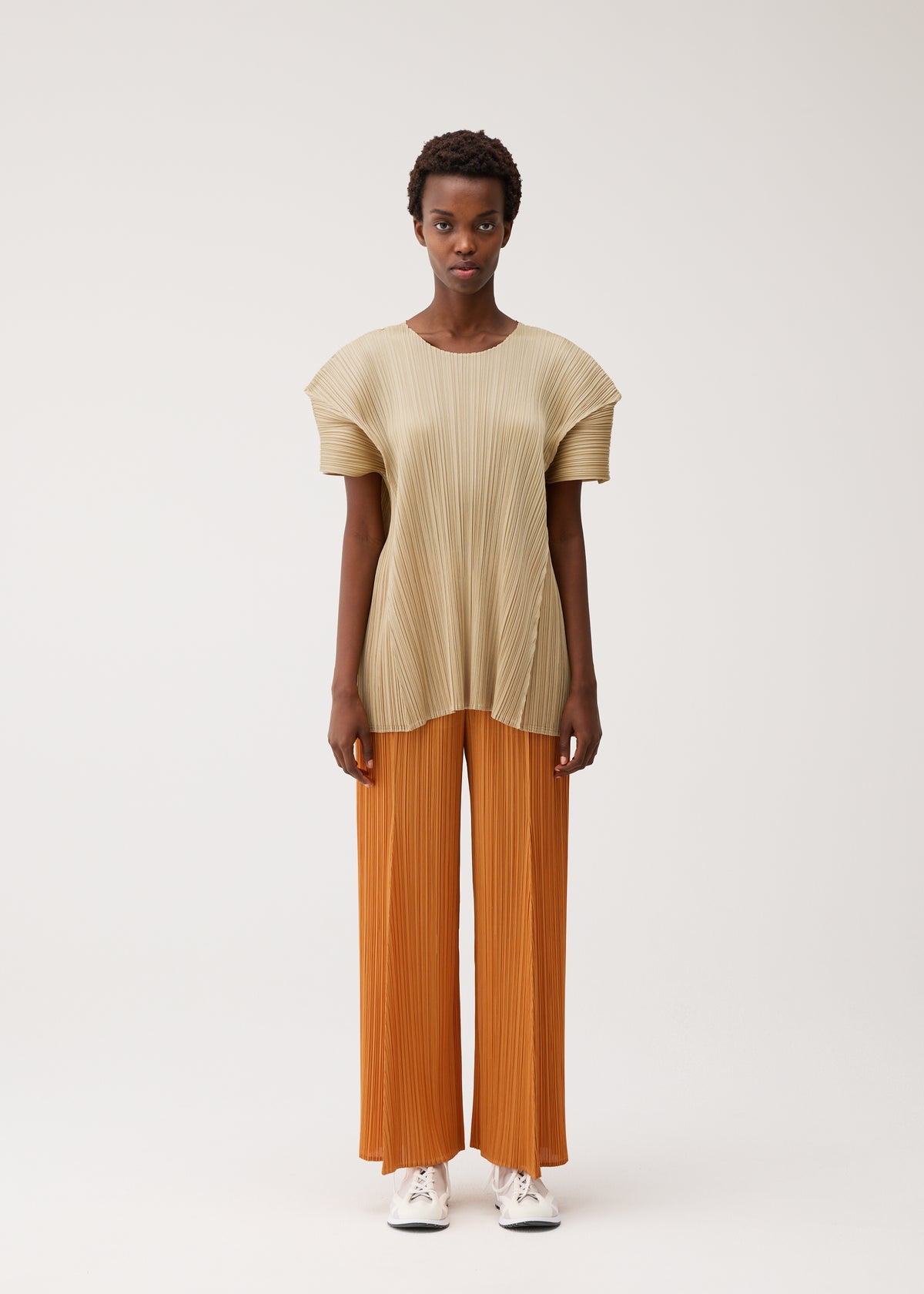 A person stands facing forward in a beige, floral-inspired pleated short-sleeved top and wide-legged orange pleated pants from BOUQUET by PLEATS PLEASE ISSEY MIYAKE, paired with white sneakers against a plain light background. view 5 of 9