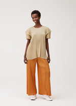A person smiles in the BOUQUET top and wide orange pleated pants from PLEATS PLEASE ISSEY MIYAKE, paired with white sneakers, set against a plain light background.