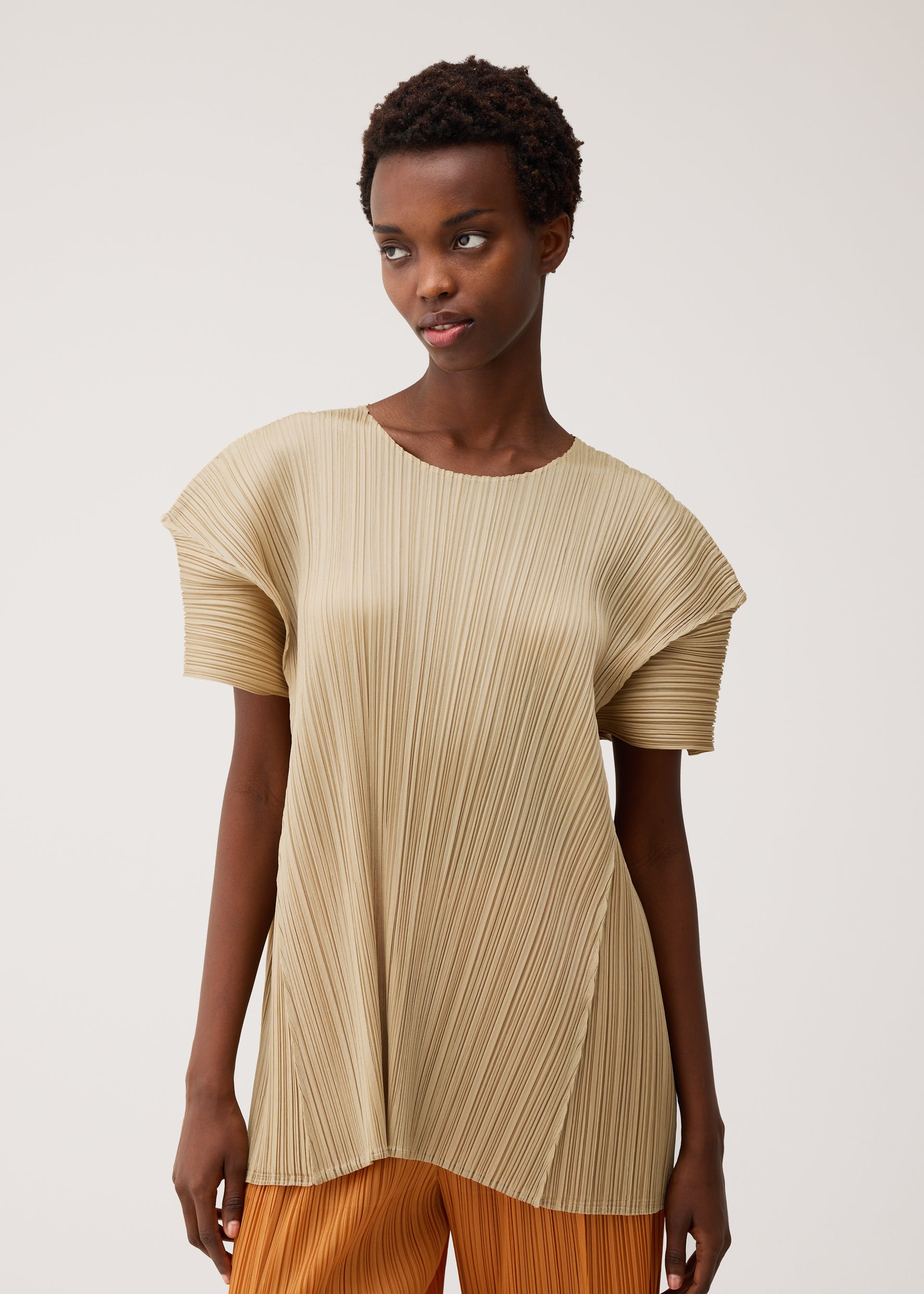 Zoomed view A person wears the beige, floral-inspired BOUQUET top by PLEATS PLEASE ISSEY MIYAKE with orange pants, standing against a plain light background and looking slightly to the side with a neutral expression.