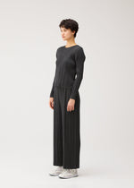 A person stands in a studio wearing the COTTON DENIM BLACK set by PLEATS PLEASE ISSEY MIYAKE, featuring a charcoal gray round-neck top and wide-leg pants paired with white shoes against a light background.