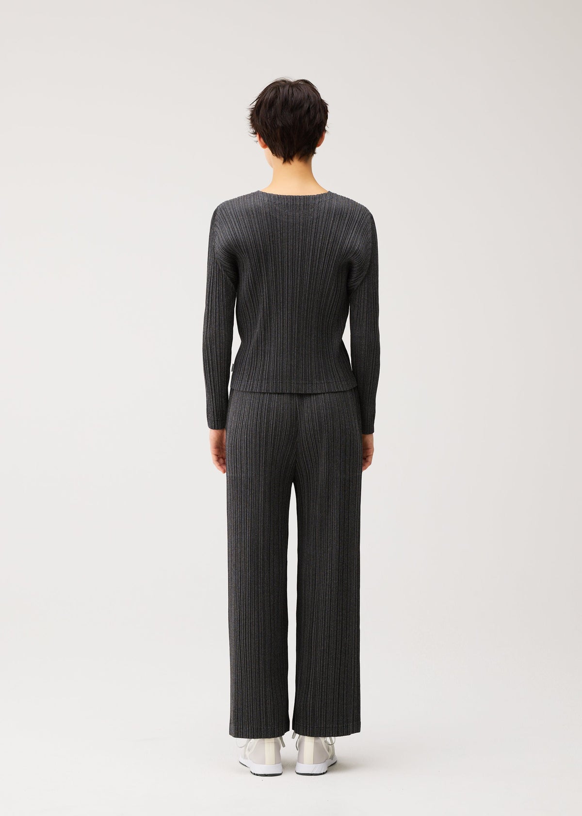 A person with short dark hair faces away, wearing the COTTON DENIM BLACK set by PLEATS PLEASE ISSEY MIYAKE and white sneakers against a plain light background. view 5 of 6