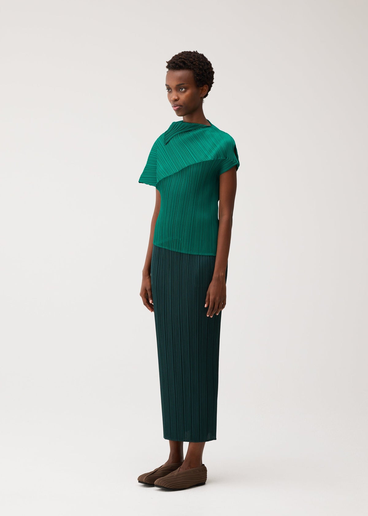 A person wears the LEAP set by PLEATS PLEASE ISSEY MIYAKE: a dark green, short-sleeved pleated top with an asymmetrical silhouette, matching long skirt, and brown shoes against a plain background. view 6 of 8