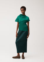 A woman poses against a plain light background wearing the LEAP green short-sleeved pleated top with collar and dark green long skirt by PLEATS PLEASE ISSEY MIYAKE, paired with brown flats.