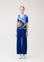A person with light skin and red hair faces forward, wearing the ROLL top and pleated blue pants by PLEATS PLEASE ISSEY MIYAKE, styled with white sneakers, standing against a light background.