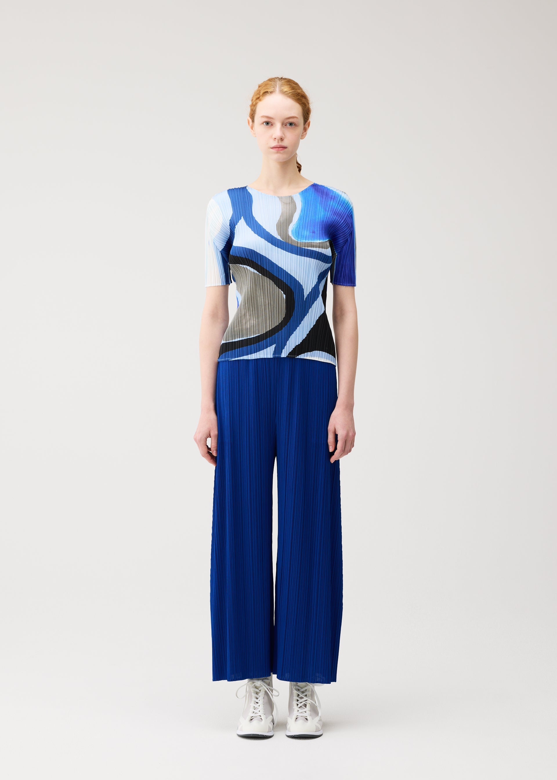 Zoomed view A person with light skin and red hair faces forward, wearing the ROLL top and pleated blue pants by PLEATS PLEASE ISSEY MIYAKE, styled with white sneakers, standing against a light background.