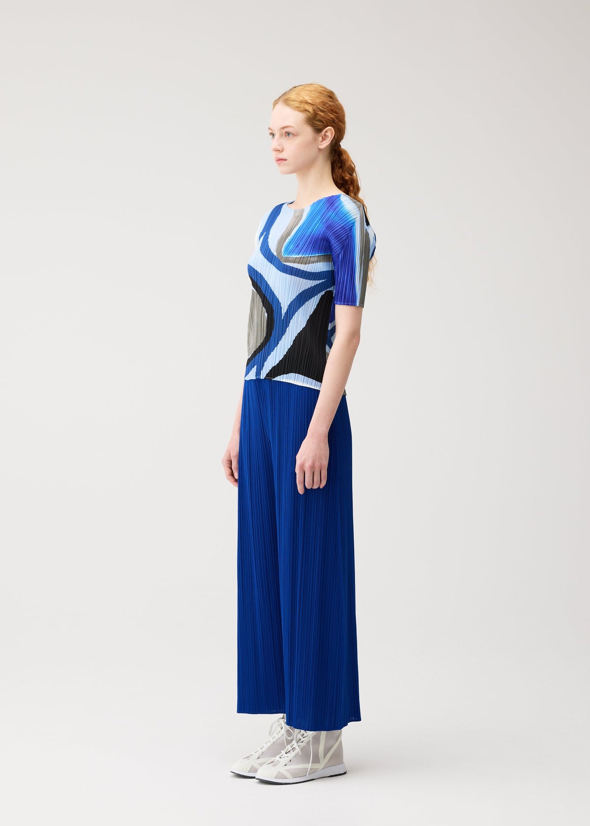A woman with long red hair in a ponytail stands sideways, wearing the ROLL blue and black round-neck printed top and wide blue pleated pants by PLEATS PLEASE ISSEY MIYAKE, paired with white lace-up shoes against a plain light background. view 5 of 7