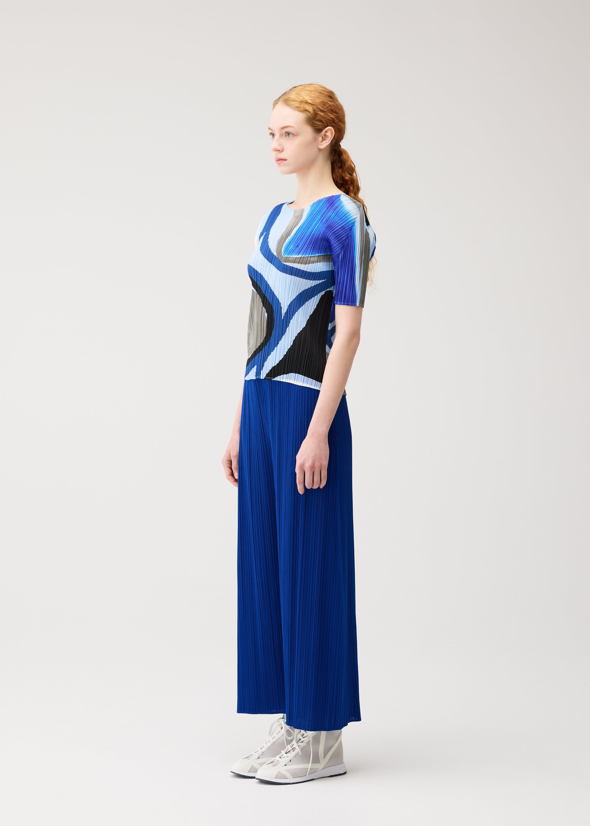 Zoomed view A woman with long red hair in a ponytail stands sideways, wearing the ROLL blue and black round-neck printed top and wide blue pleated pants by PLEATS PLEASE ISSEY MIYAKE, paired with white lace-up shoes against a plain light background.