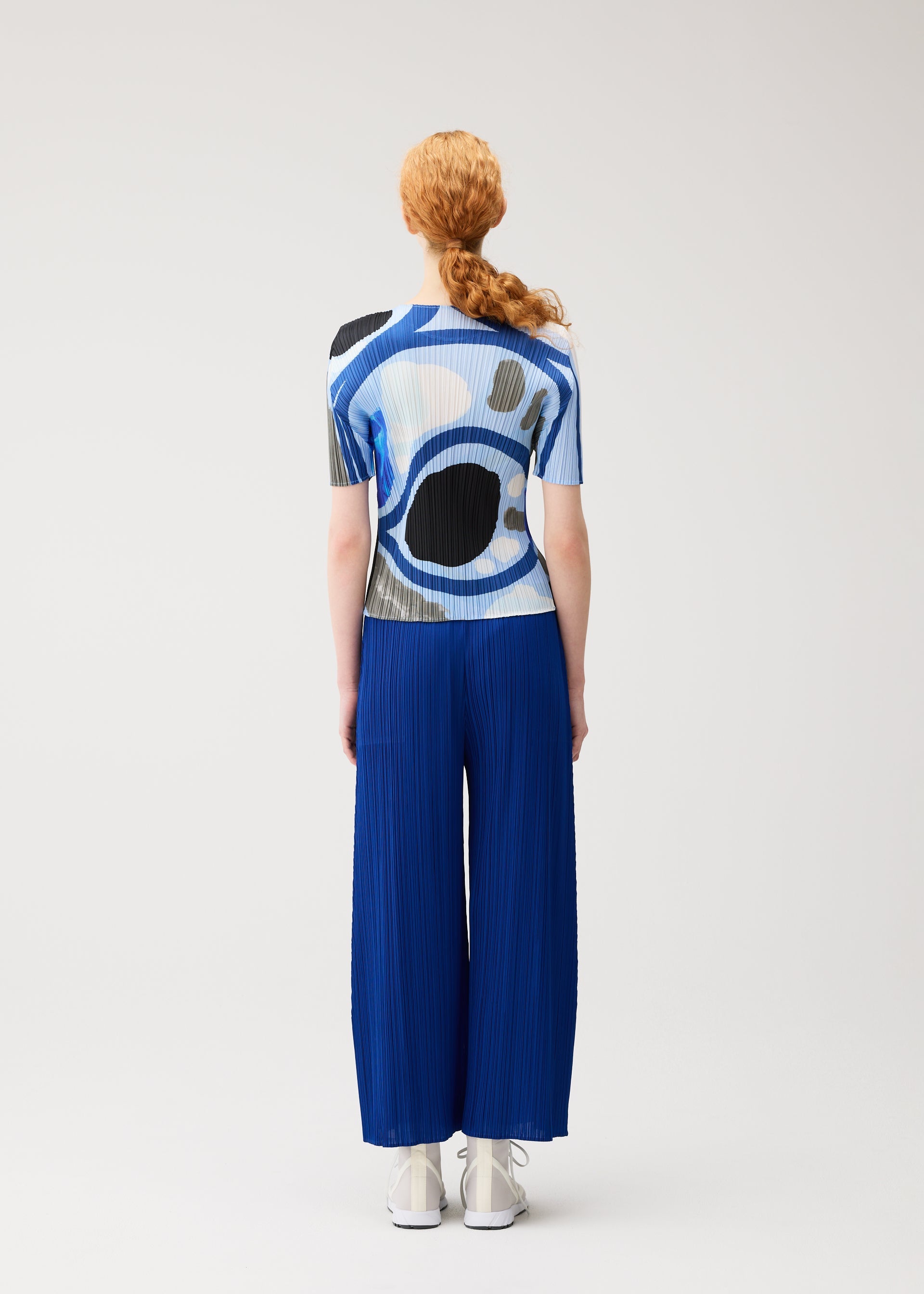 Zoomed view A person with long red hair in a braid models the ROLL printed top and blue pleated wide-leg pants by PLEATS PLEASE ISSEY MIYAKE, standing facing away against a plain light background.