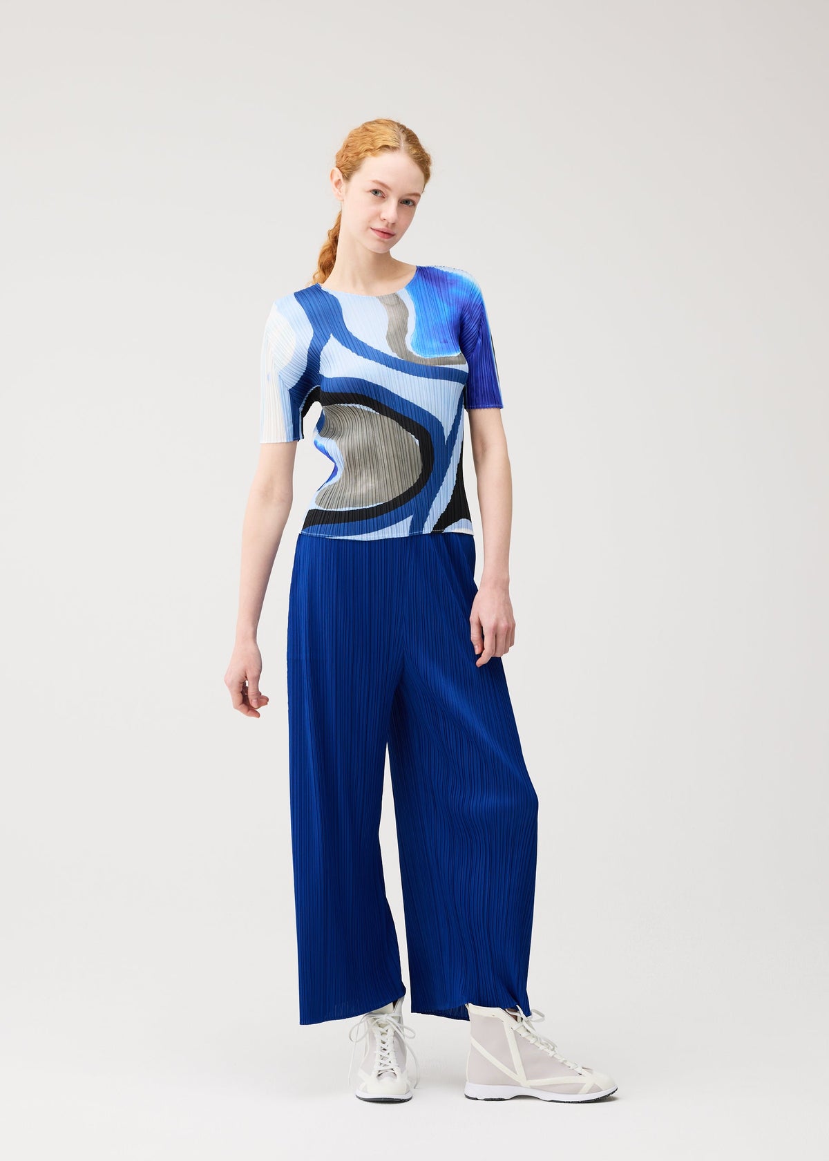 A woman with red hair in a ponytail wears the ROLL top and wide blue pleated pants by PLEATS PLEASE ISSEY MIYAKE, paired with white sneakers, standing against a plain light background. view 7 of 7
