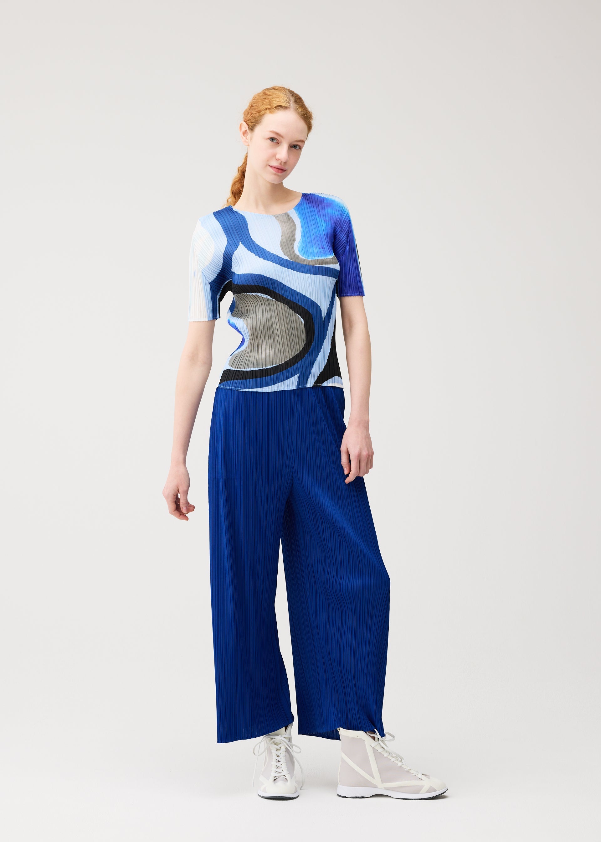 Zoomed view A woman with red hair in a ponytail wears the ROLL top and wide blue pleated pants by PLEATS PLEASE ISSEY MIYAKE, paired with white sneakers, standing against a plain light background.