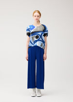 A person with light skin and reddish hair tied back stands against a plain background, wearing PLEATS PLEASE ISSEY MIYAKE's ROLL sushi print round-neck top and pleated wide-leg blue pants, paired with white lace-up shoes.