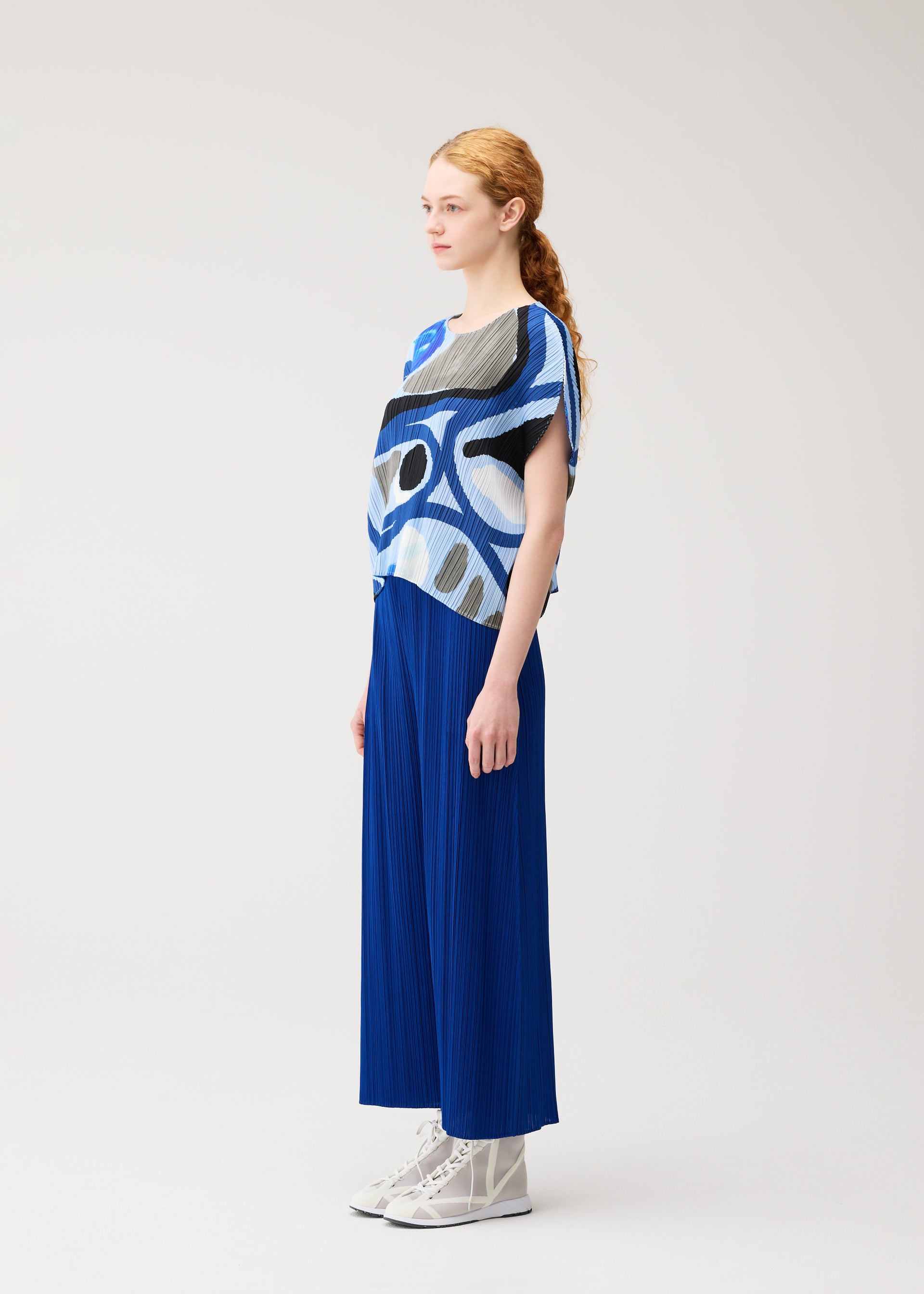 Zoomed view A woman with red hair in a ponytail stands sideways, wearing the ROLL round-neck top and pleated blue skirt from PLEATS PLEASE ISSEY MIYAKE, paired with white lace-up shoes against a plain light background.