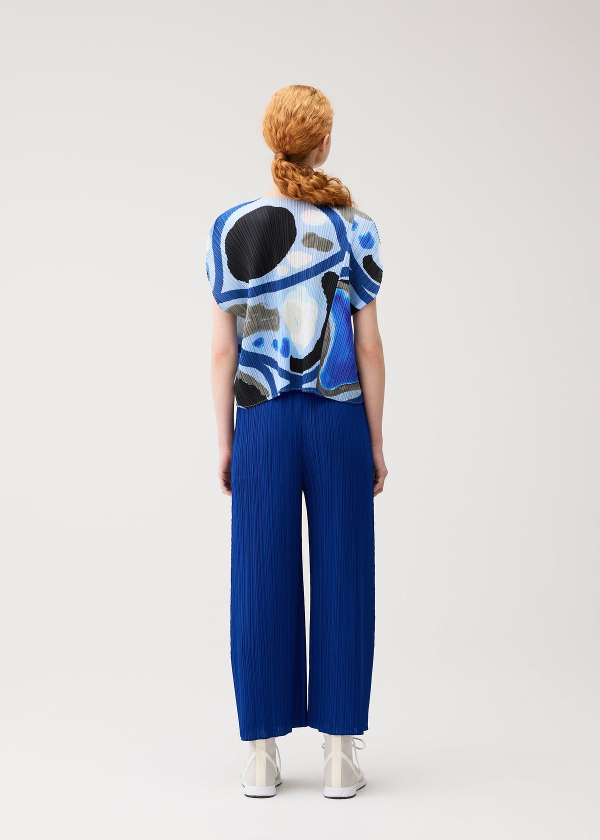 A person with long, reddish hair in a braid faces away from the camera, wearing PLEATS PLEASE ISSEY MIYAKE's ROLL top with blue and black abstract patterns and bright blue pleated wide-leg pants against a plain light background. view 7 of 8