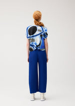 A person with long, reddish hair in a braid faces away from the camera, wearing PLEATS PLEASE ISSEY MIYAKE's ROLL top with blue and black abstract patterns and bright blue pleated wide-leg pants against a plain light background.