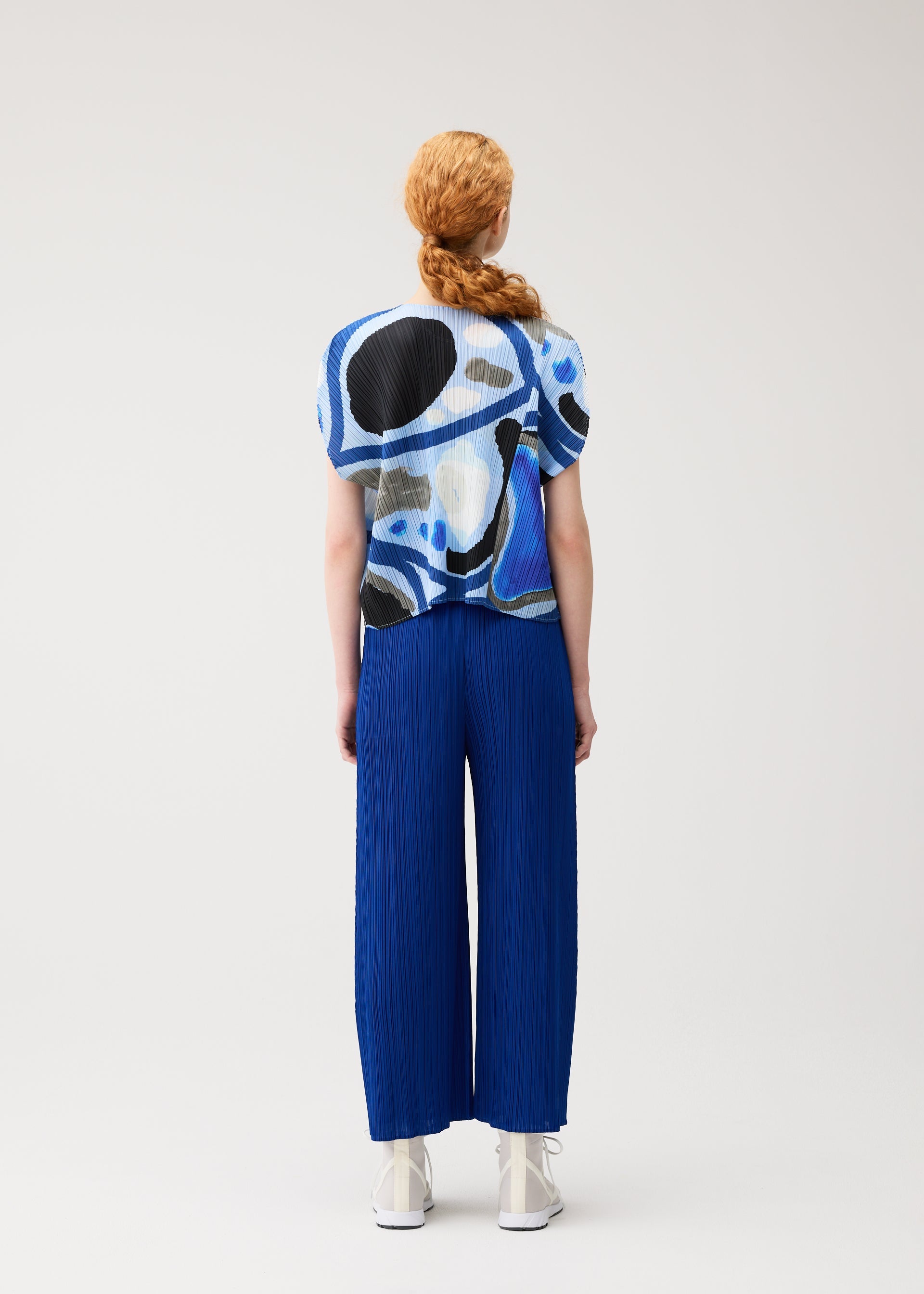 Zoomed view A person with long, reddish hair in a braid faces away from the camera, wearing PLEATS PLEASE ISSEY MIYAKE's ROLL top with blue and black abstract patterns and bright blue pleated wide-leg pants against a plain light background.