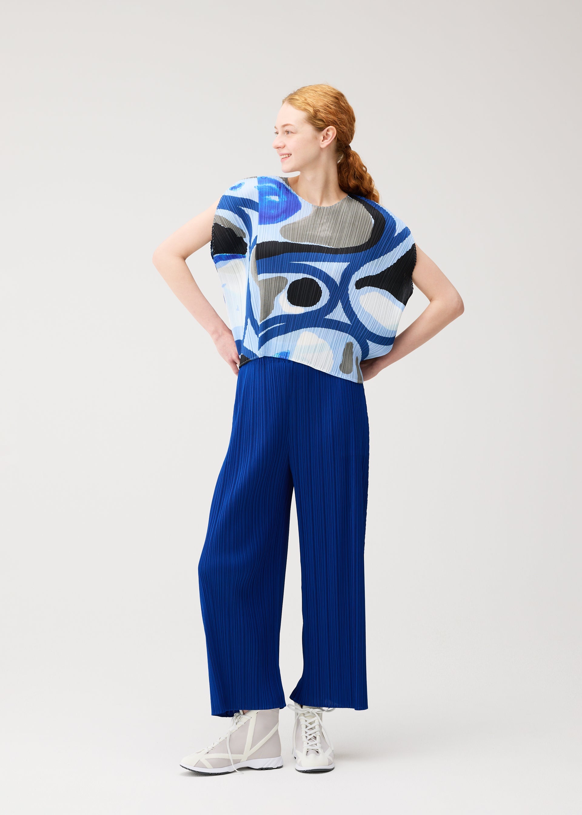 Zoomed view A red-haired woman in a ponytail wears the ROLL top and wide pleated pants by PLEATS PLEASE ISSEY MIYAKE, featuring blue, black, and white patterns, paired with white lace-up shoes, smiling against a light background.