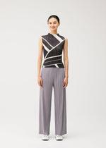 A woman stands against a white background, smiling with her arms relaxed at her sides, wearing the FOLD LINE high-neck sleeveless black top adorned with striking diagonal white stripes and wide-legged gray pleated pants by PLEATS PLEASE ISSEY MIYAKE, paired with white sneakers.
