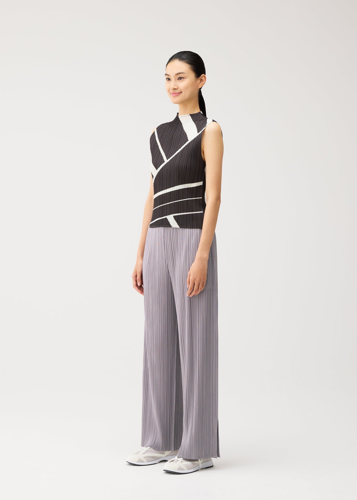 A woman with a ponytail models the FOLD LINE high-neck, sleeveless top by PLEATS PLEASE ISSEY MIYAKE, which features a black and white diagonal wrapping pattern. She pairs it with wide-leg gray pleated pants and white sneakers, standing against a plain light background. view 5 of 7