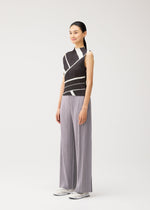 A woman with a ponytail models the FOLD LINE high-neck, sleeveless top by PLEATS PLEASE ISSEY MIYAKE, which features a black and white diagonal wrapping pattern. She pairs it with wide-leg gray pleated pants and white sneakers, standing against a plain light background.
