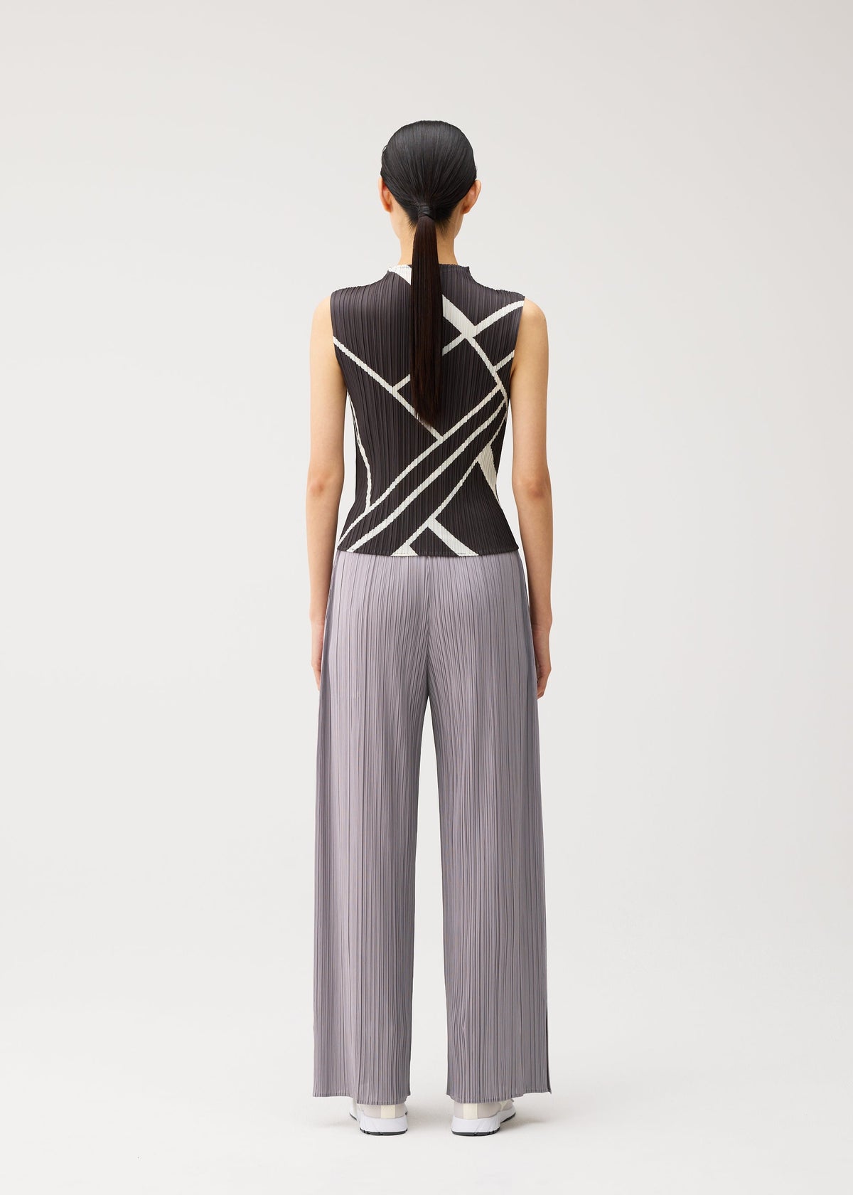 A person stands with their back to the camera, wearing the FOLD LINE high-neck sleeveless black top featuring a white diagonal wrapping pattern and wide-legged, light gray pleated pants from PLEATS PLEASE ISSEY MIYAKE. Their hair is straight and tied back in a low ponytail. The plain white background further highlights the minimalist design. view 6 of 7