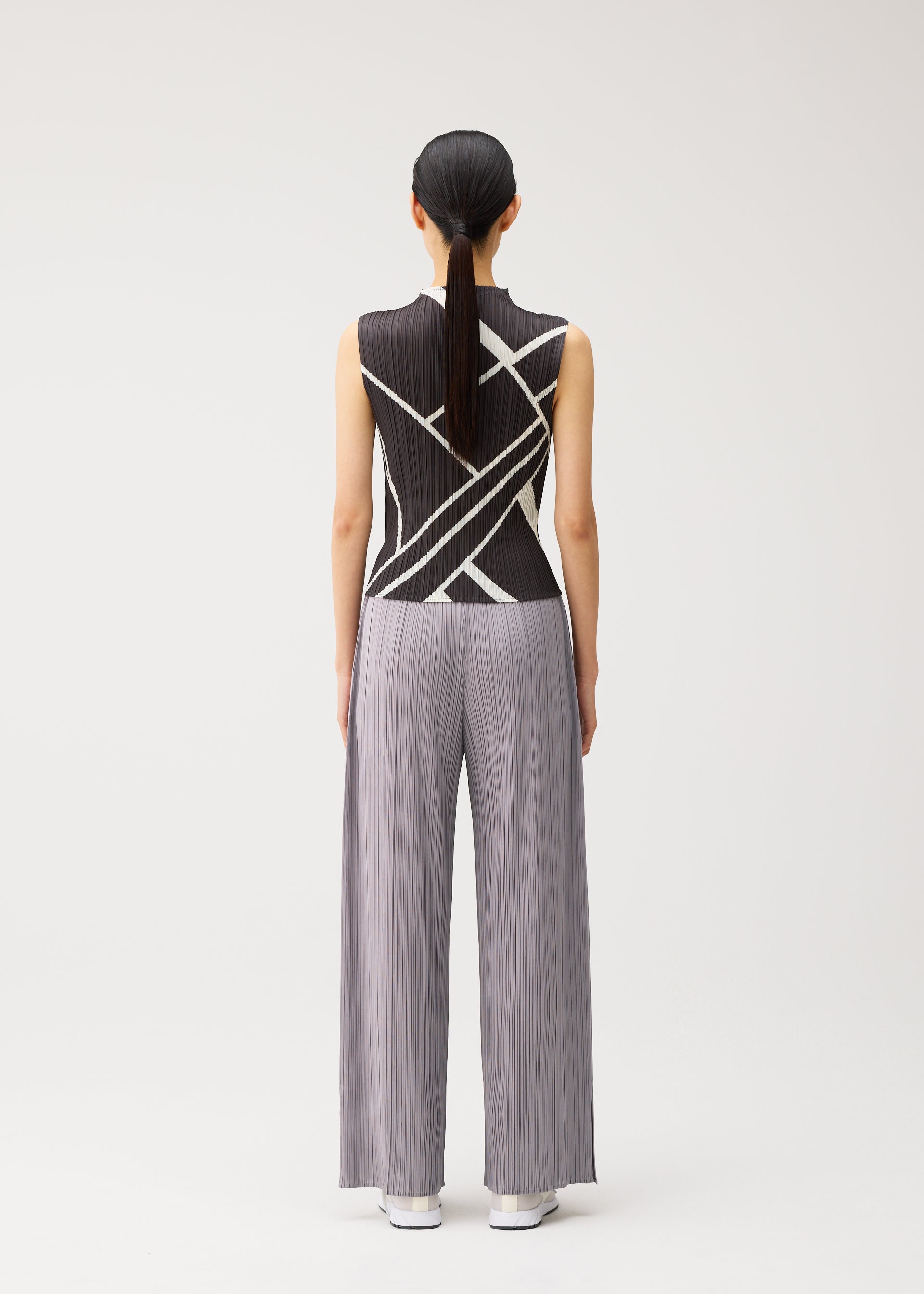 Zoomed view A person stands with their back to the camera, wearing the FOLD LINE high-neck sleeveless black top featuring a white diagonal wrapping pattern and wide-legged, light gray pleated pants from PLEATS PLEASE ISSEY MIYAKE. Their hair is straight and tied back in a low ponytail. The plain white background further highlights the minimalist design.