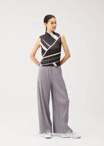 A woman stands against a plain background wearing the FOLD LINE high-neck sleeveless top by PLEATS PLEASE ISSEY MIYAKE, featuring a black and white geometric diagonal wrapping pattern. She pairs it with loose gray pleated pants and white sneakers, her hair pulled back and her hands behind her back.