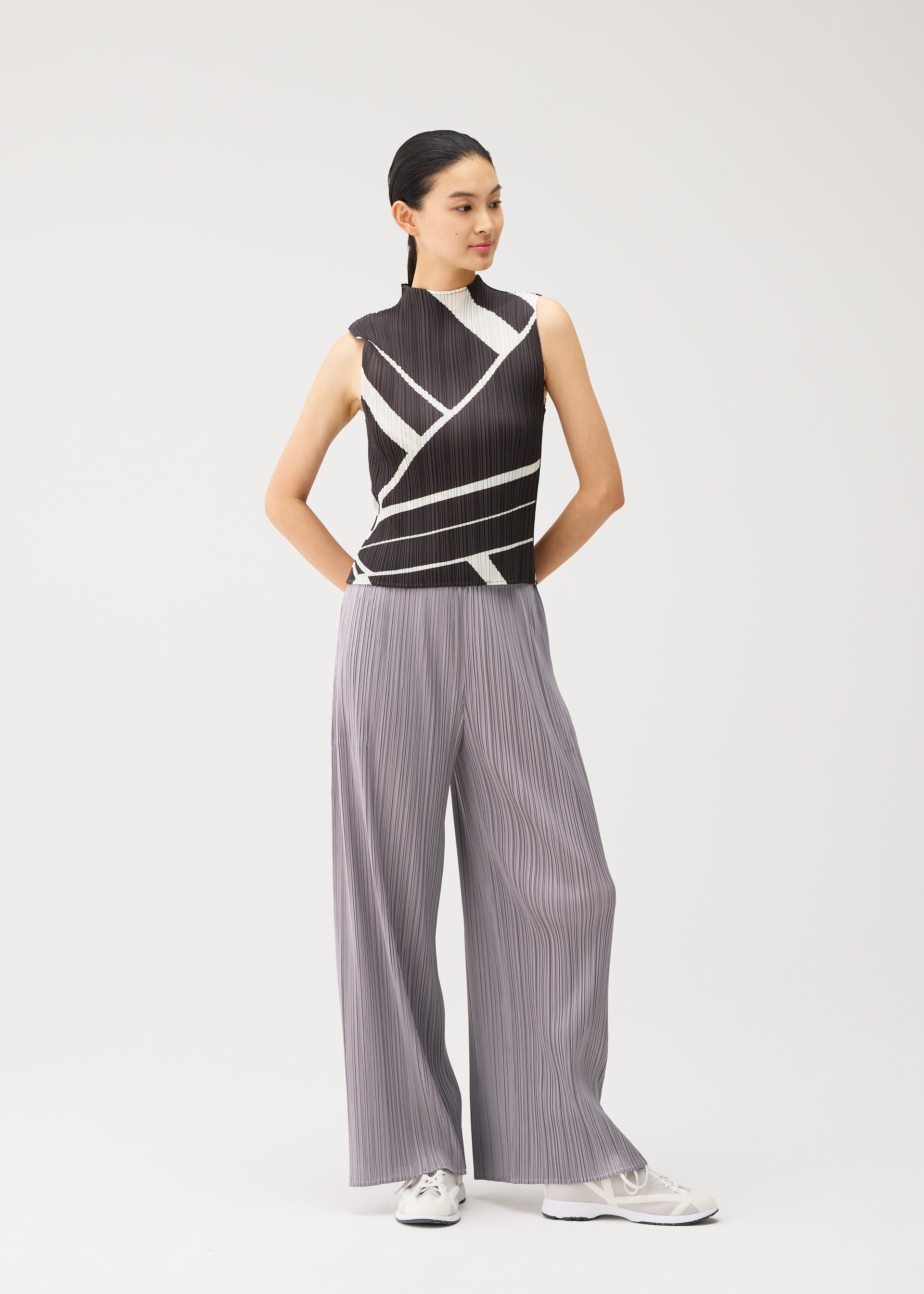 Zoomed view A woman stands against a plain background wearing the FOLD LINE high-neck sleeveless top by PLEATS PLEASE ISSEY MIYAKE, featuring a black and white geometric diagonal wrapping pattern. She pairs it with loose gray pleated pants and white sneakers, her hair pulled back and her hands behind her back.