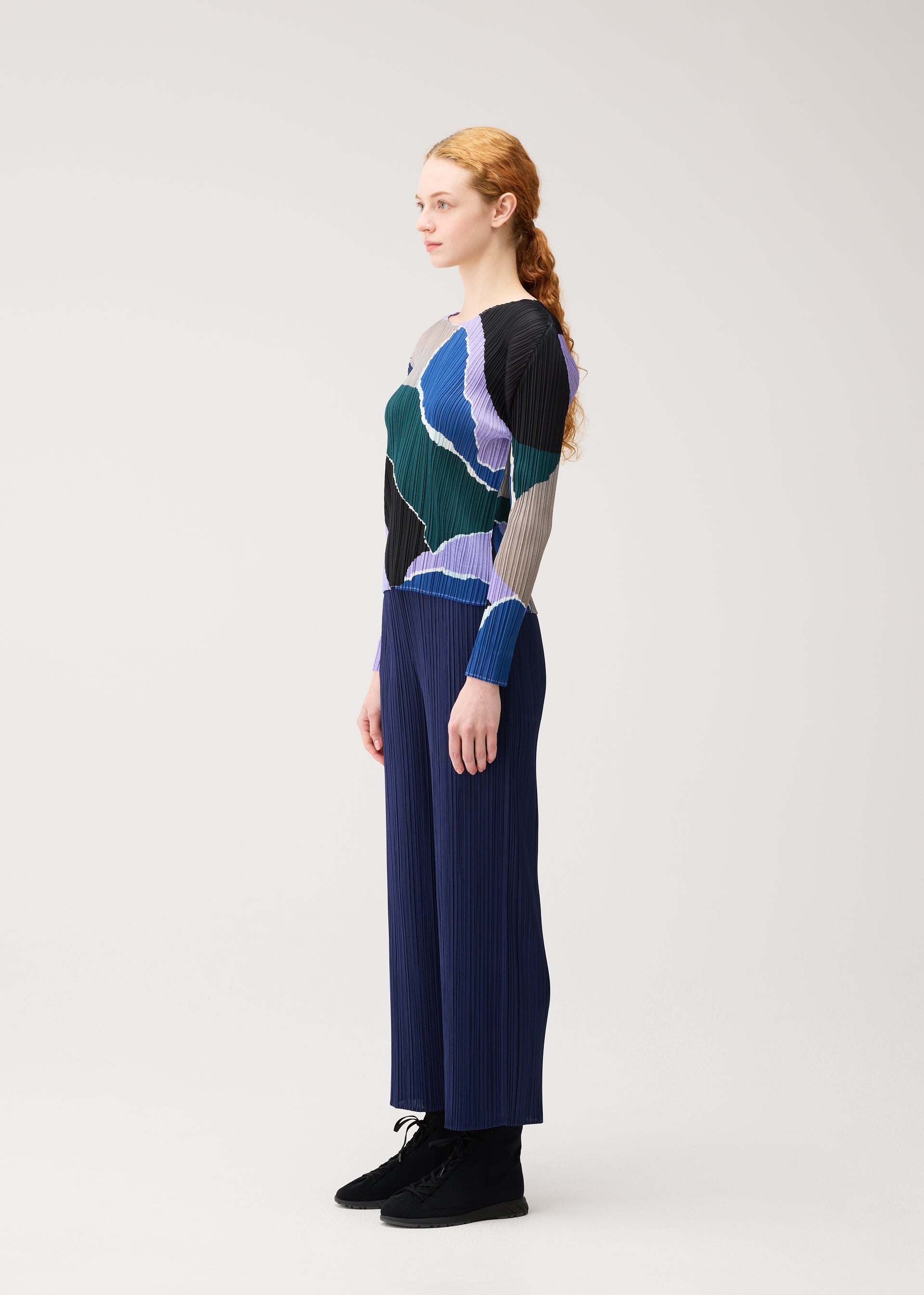 Zoomed view A person with long curly hair stands in profile against a plain light background, wearing the PLEATS PLEASE ISSEY MIYAKE “SURPRISE!” shirt featuring a colorful unique pattern, paired with blue pleated pants and black shoes.
