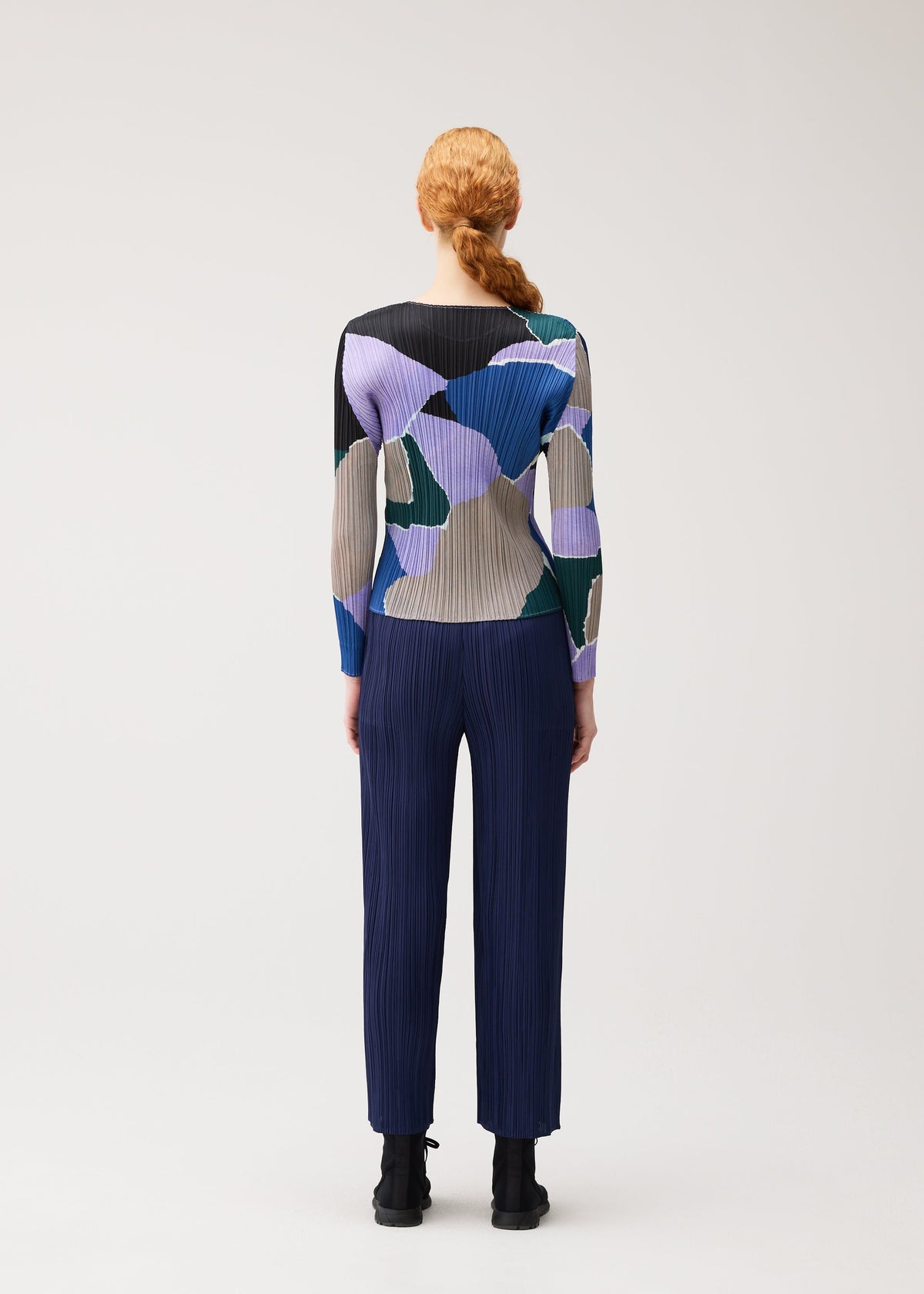 A person with red hair in a braid, wearing the SURPRISE! round-neck long-sleeved top with a colorful pattern and dark pleated pants by PLEATS PLEASE ISSEY MIYAKE, stands facing away against a plain light background. view 6 of 7