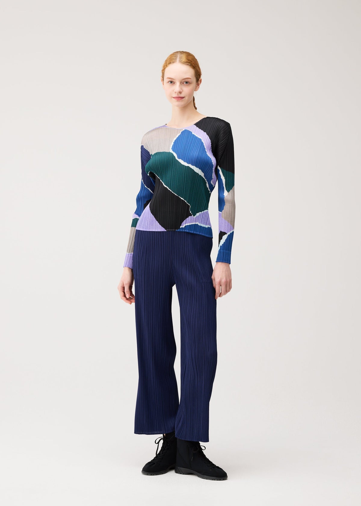 A person stands against a plain light background wearing the PLEATS PLEASE ISSEY MIYAKE SURPRISE! round-neck long-sleeved top featuring a colorful, abstract pattern, paired with blue pleated pants and black shoes. view 7 of 7