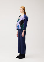 A person with light skin and red hair in a ponytail wears the SURPRISE! blue pleated outfit by PLEATS PLEASE ISSEY MIYAKE, featuring a boat-neck top with a colorful pattern and black shoes, standing against a plain light background.
